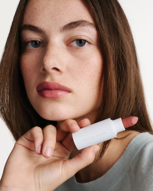Woman holding a white lip balm with visible brand name against a neutral background
