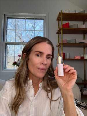 Woman holding a skincare product in a room with a window and shelves.