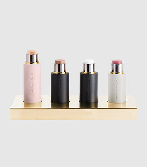Four makeup sticks in a gold tray against a grey background.